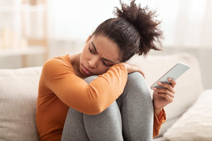 beautiful young Black woman sitting on couch with her knees drawn to her chest, resting her head while holding a cell phone in one hand - women and substance abuse
