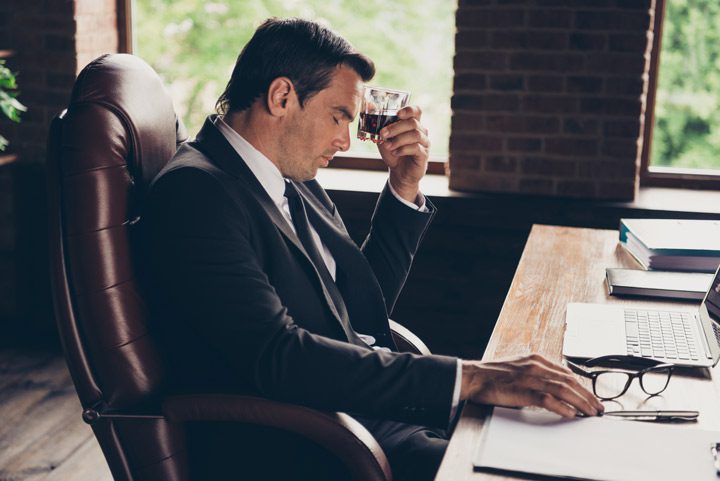 businessman in suit at his office desk looking stressed and holding a tumbler of liquor up to his forehead - addiction and work
