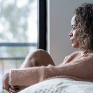 Woman sitting by a window with a thoughtful, serious expression, hugging her knees