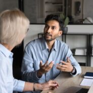 Man and woman sitting at a desk having a serious discussion in an office setting, with a laptop, notebook, and papers on the table.