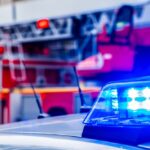 Close-up of a police car with flashing blue lights, with a fire truck blurred in the background, symbolizing emergency response.