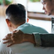 A man gently places a comforting hand on another man’s back as they sit together indoors, offering emotional support in a quiet, reflective moment.