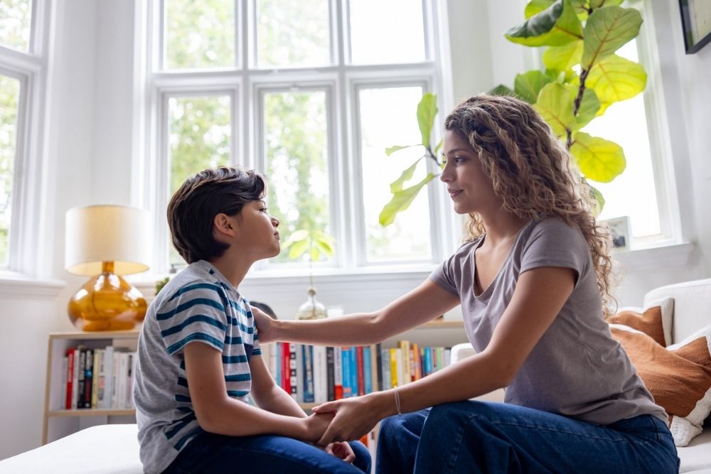 Mother sitting on a couch having a serious conversation with her young son in a bright living room with large windows and houseplants.