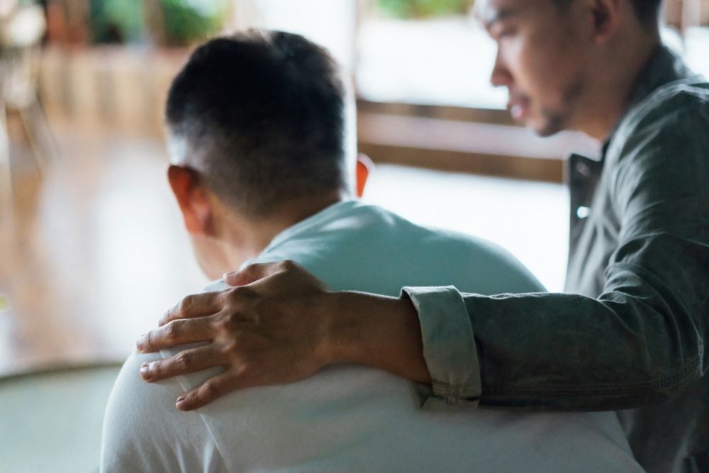 A man gently places a comforting hand on another man’s back as they sit together indoors