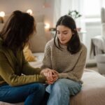 A woman and a teenage girl sit on the edge of a bed holding hands. Both look down with thoughtful, serious expressions, sharing a quiet moment in a softly lit room. Ask ChatGPT