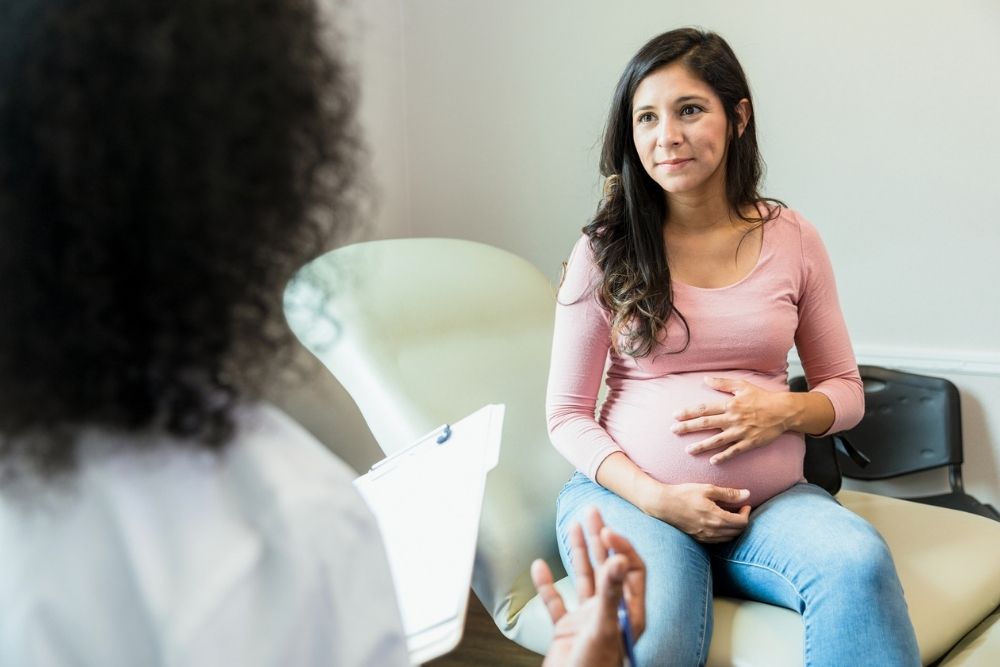 A pregnant woman sitting in a chair, holding her belly, listening to a healthcare professional during a consultation.