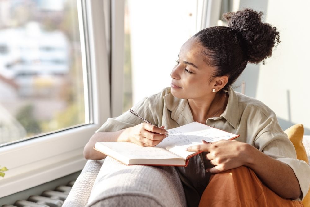 A woman sits by a window holding a notebook and pen, looking outside thoughtfully while writing.