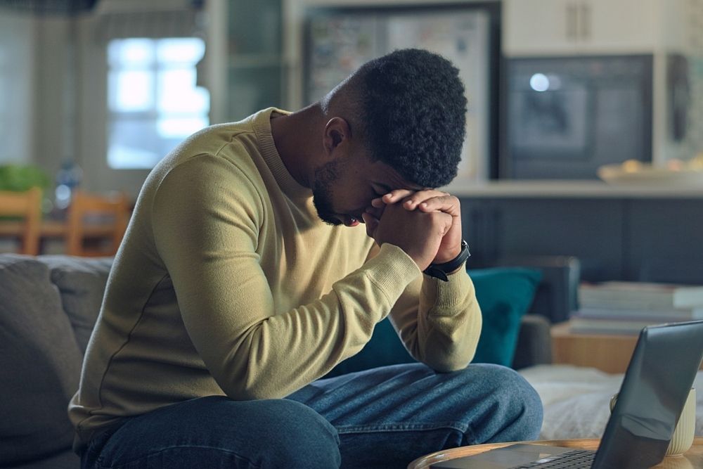 A man sits on a couch with his head bowed and hands clasped in front of a laptop, appearing stressed or deep in thought.