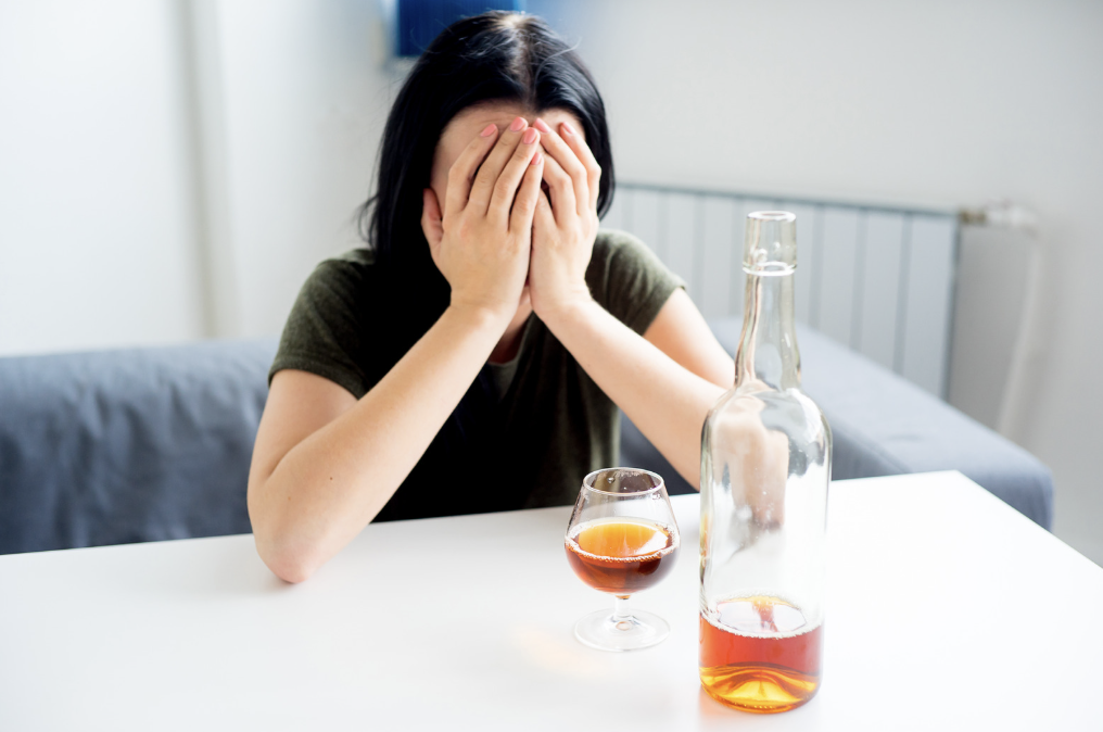 A person with their face covered sitting in front of alcohol.