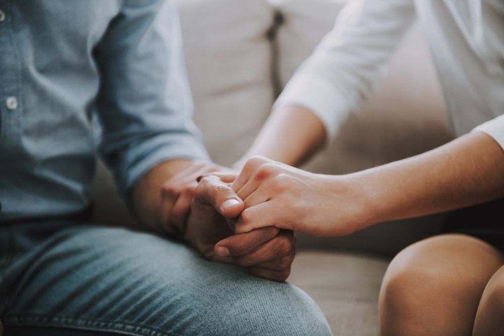 Two people sit close together on a couch, holding hands in a supportive gesture that conveys care, connection, and emotional support.