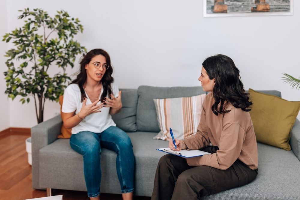 Victory - What Is Dual Diagnosis and Do You Need It. Two women sit on a gray coach. One woman in a white sirt is speaking to another in a brown shirt writing on a piece of paper and clip board.